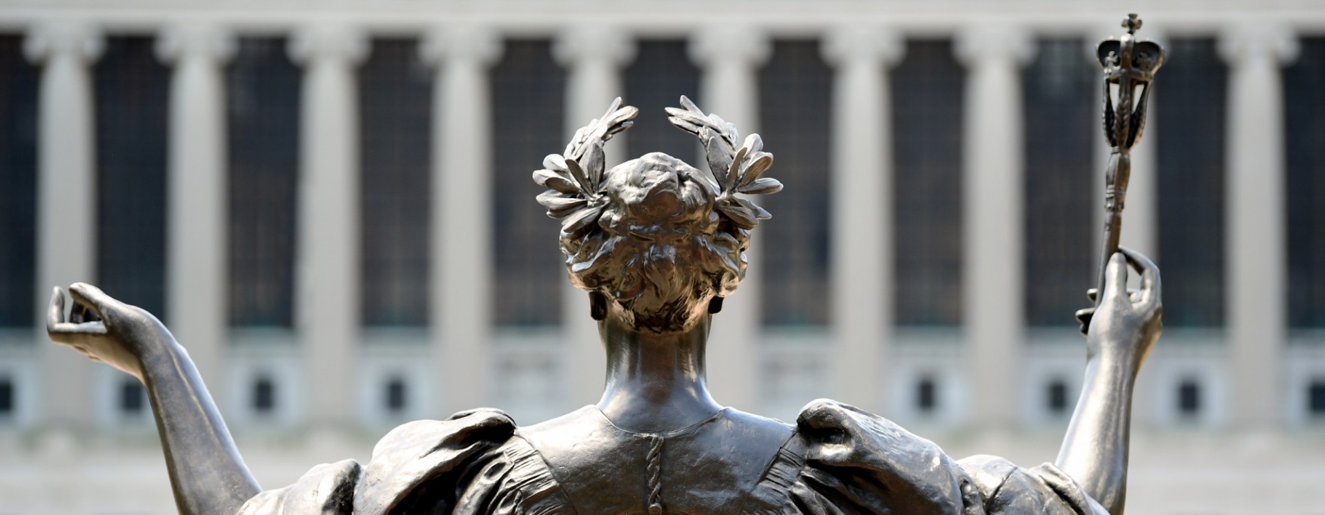 Photograph of back of Alma Matter statue's head and shoulders from the steps of Low Library looking towards a the columns of Butler Library (out of focus)